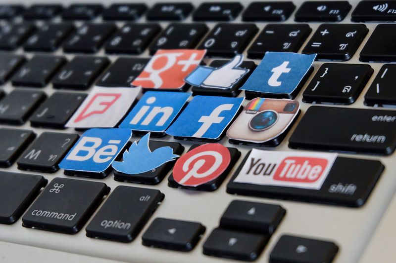 Close-up of a laptop keyboard with colorful social media icons placed on several keys (Facebook, Twitter, Instagram, LinkedIn, Pinterest, YouTube, Tumblr, Behance).