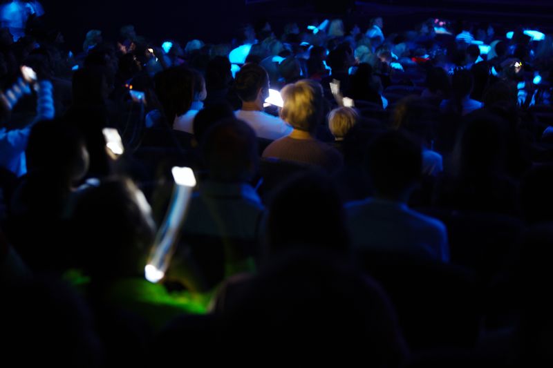Audience in a dark concert hall holding light sticks under blue stage lighting.