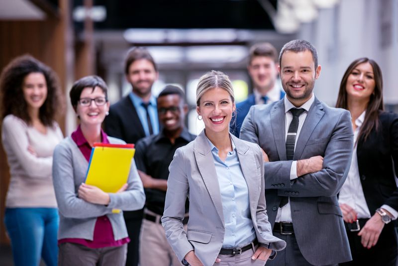 Diverse group of professionals posing in a modern office corridor, front and center a blonde woman in a light gray blazer and blue shirt.
