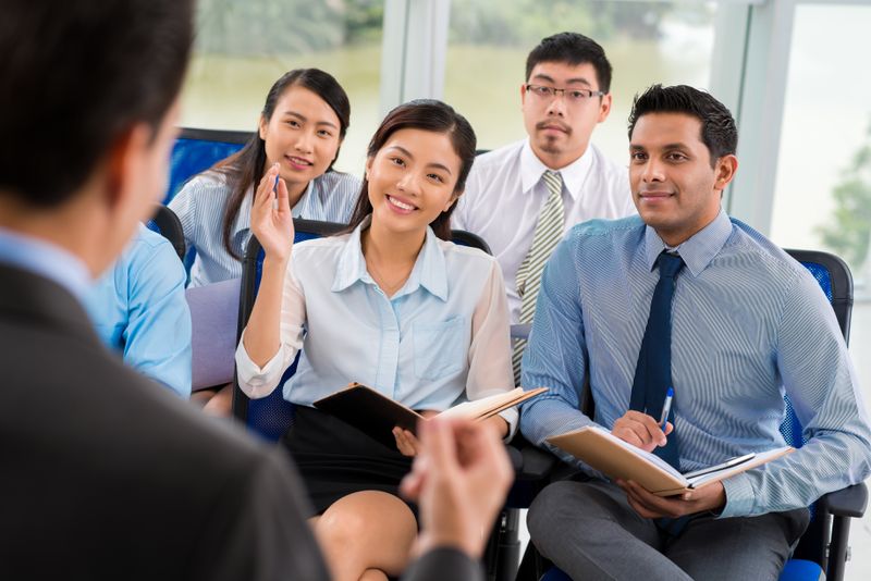 Team of four professionals in a meeting; woman in front raises her hand to contribute while others listen and take notes.