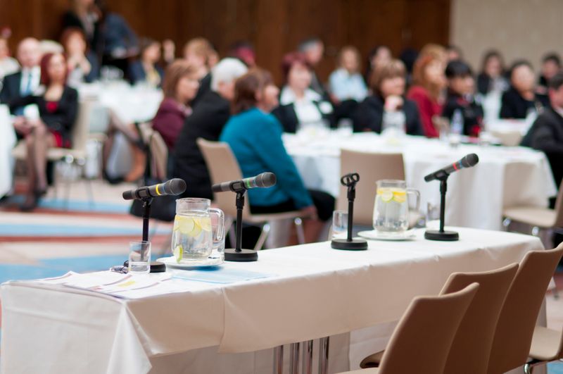 Conference room with microphones on a table ready for speakers and an audience in the background.