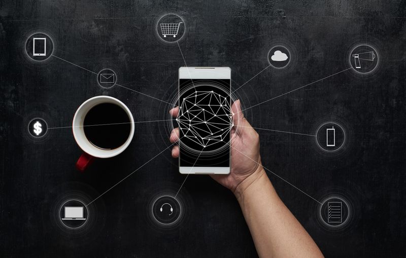 Hand holding a smartphone with network lines connecting to icons (cloud, shopping cart, money, email) around a coffee cup on a dark background.