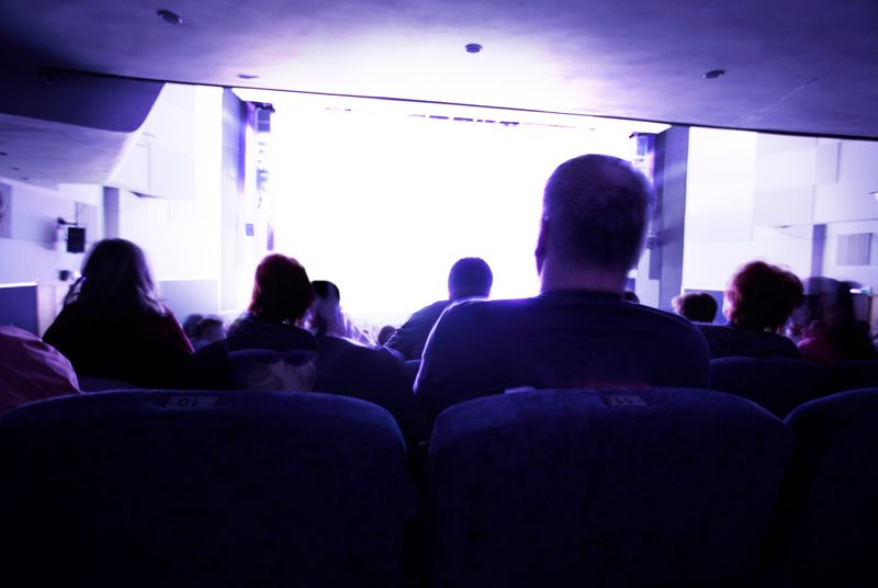 Back view of an audience seated in a dim theater, facing a bright stage.
