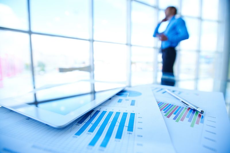 Desk with a tablet and printed charts showing blue bars and colorful graphs; a blurred person in a blue shirt stands in the background.