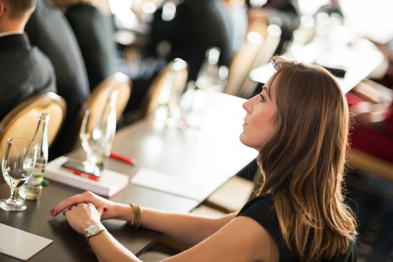 mnFC7iLYY4I_w-xETRyg5 - Prodify Digital Side profile of a woman with light brown hair listening intently at a conference table, hands resting on the desk with glassware and notebooks nearby; other attendees blurred in the background.