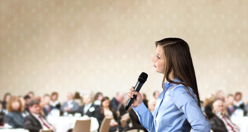 Woman in a blue blouse speaks into a handheld microphone while addressing a conference audience in a hall.